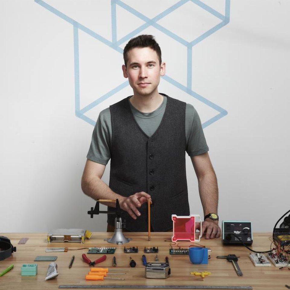 Image features man holding a pencil upright while standing in front of a table covered in tools.