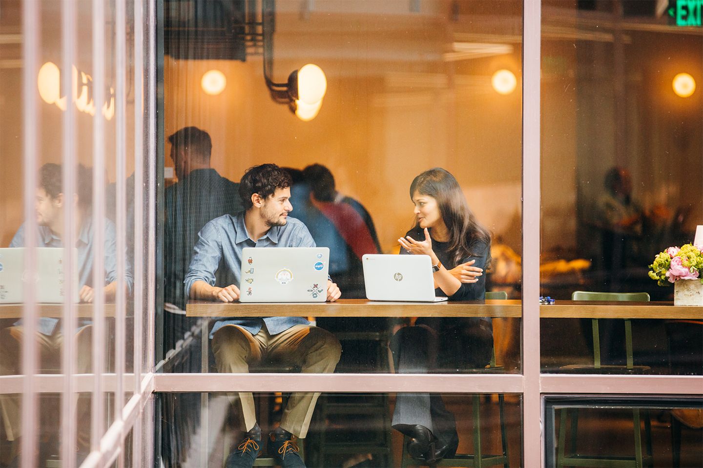 Two people working on laptops in a cafe have a conversation.