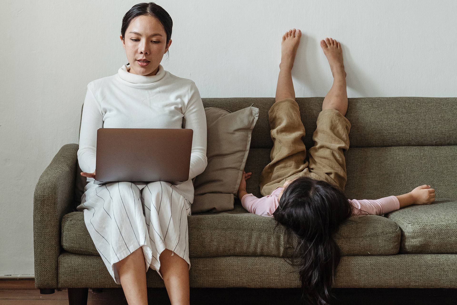 A person working from home on a laptop with their kid lying on the sofa next to them.