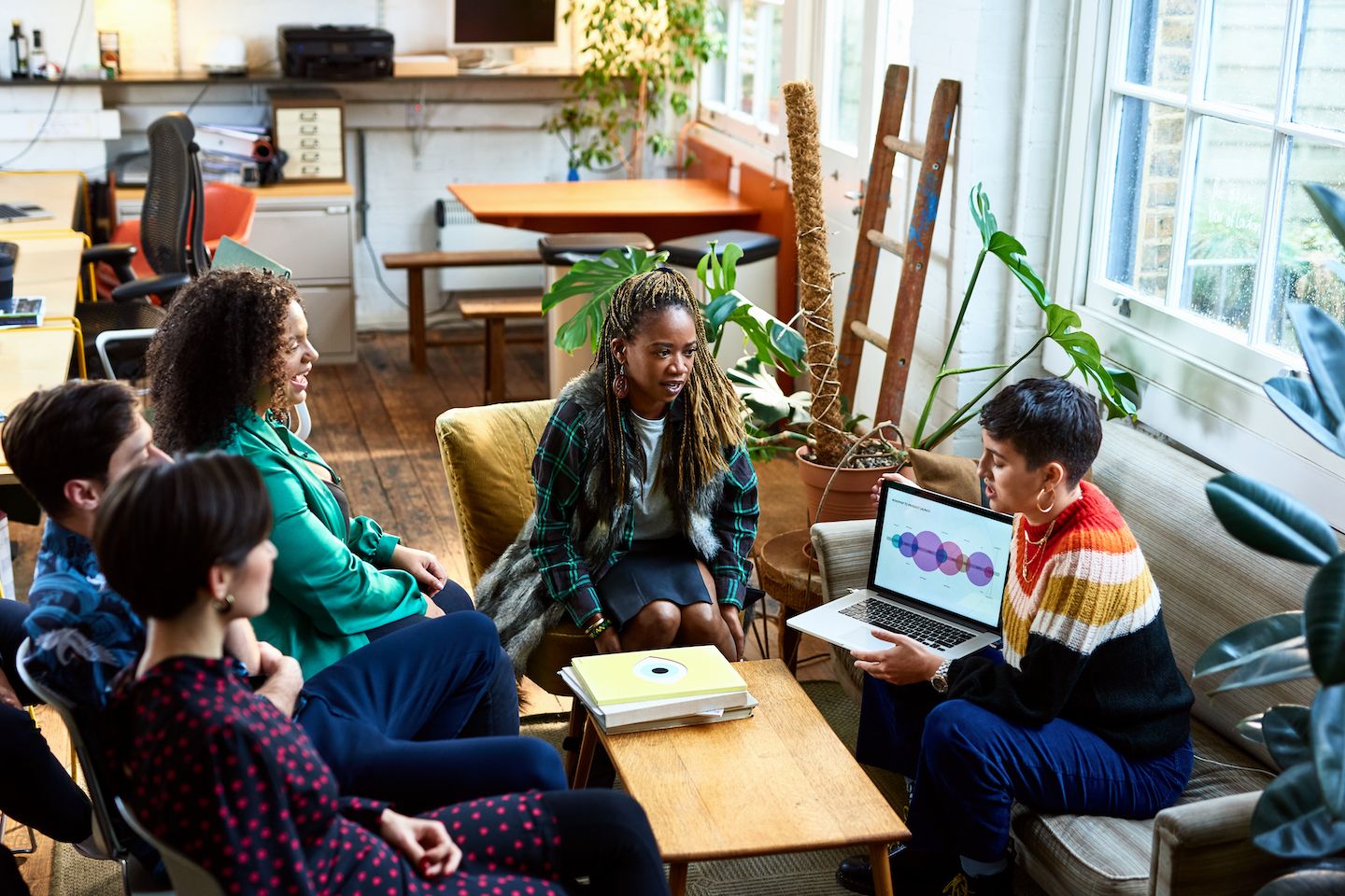 A group of people sit in an office breakout area discussing an issue around a laptop.