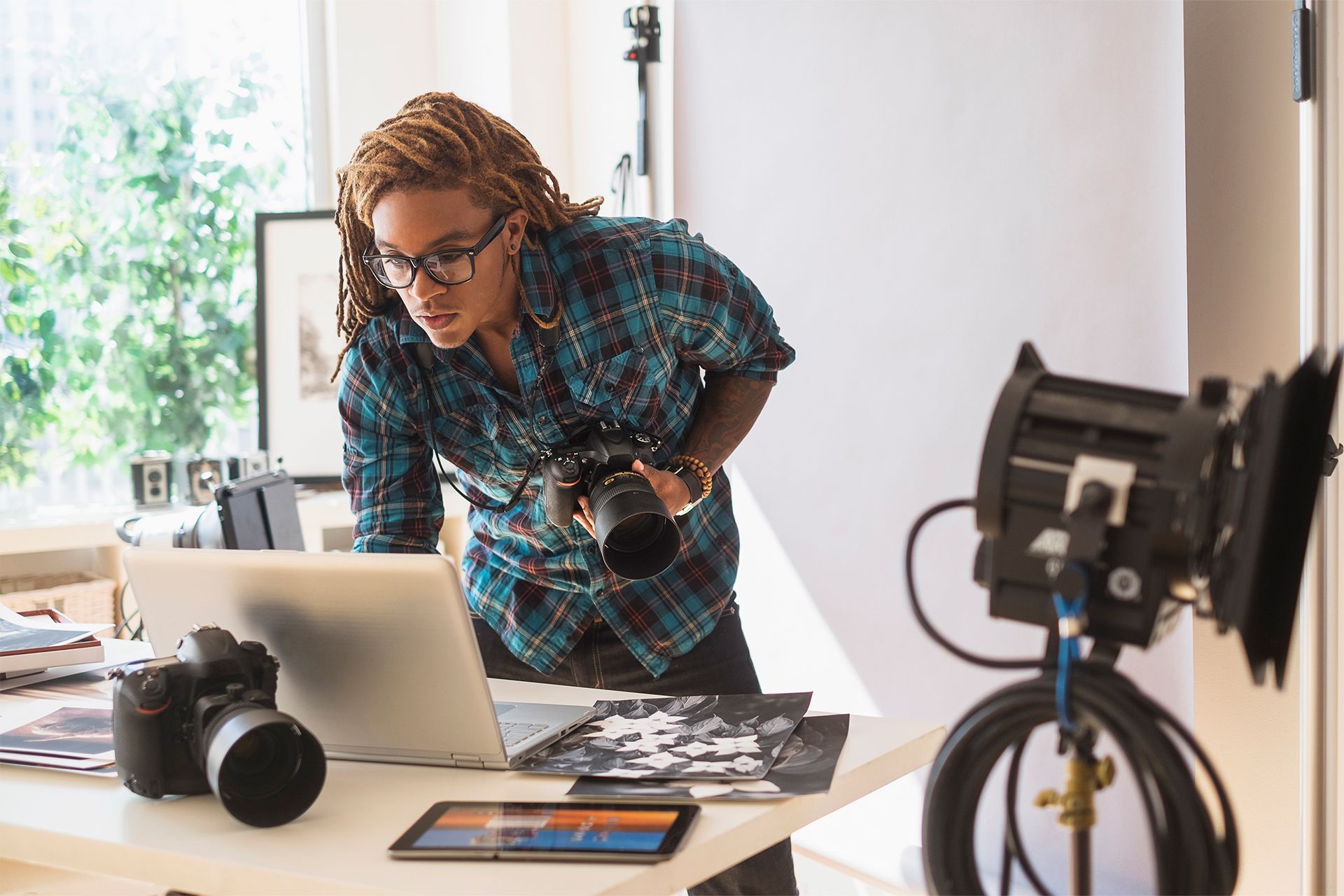 A person in a photography studio leans over their laptop while holding a camera.