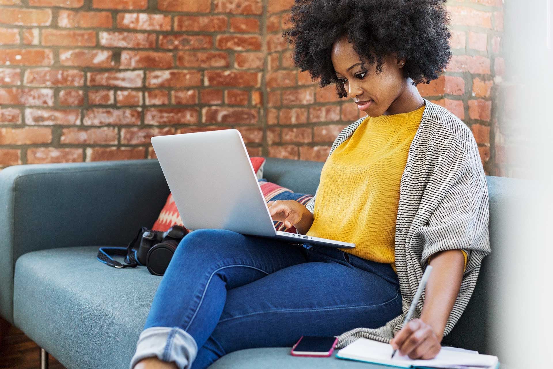 A person sitting on a couch looking at a laptop screen, writing in their notebook on the couch.