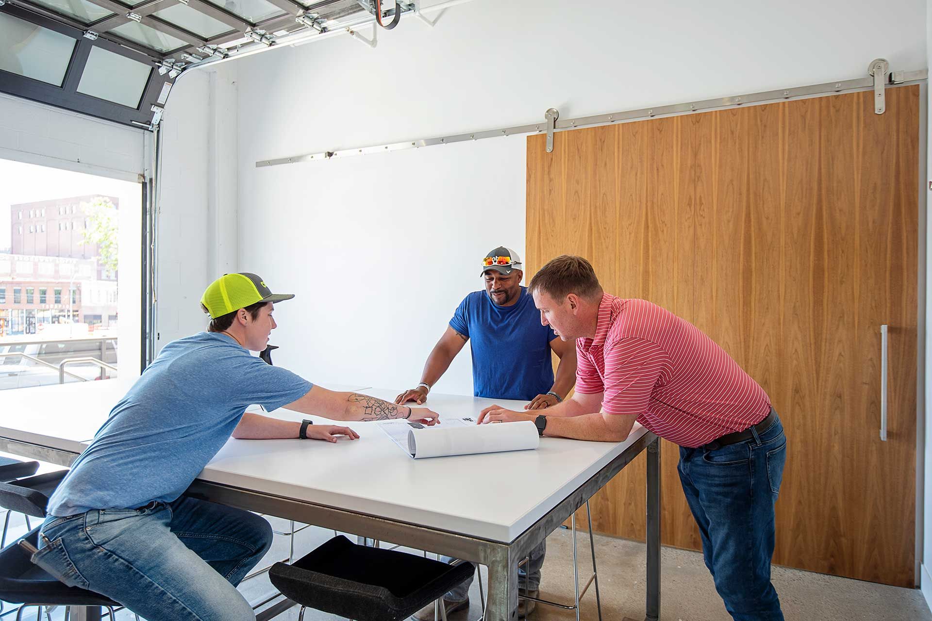 Drei Männer betrachten Pläne auf einem Schreibtisch neben einem offenen Garagentor