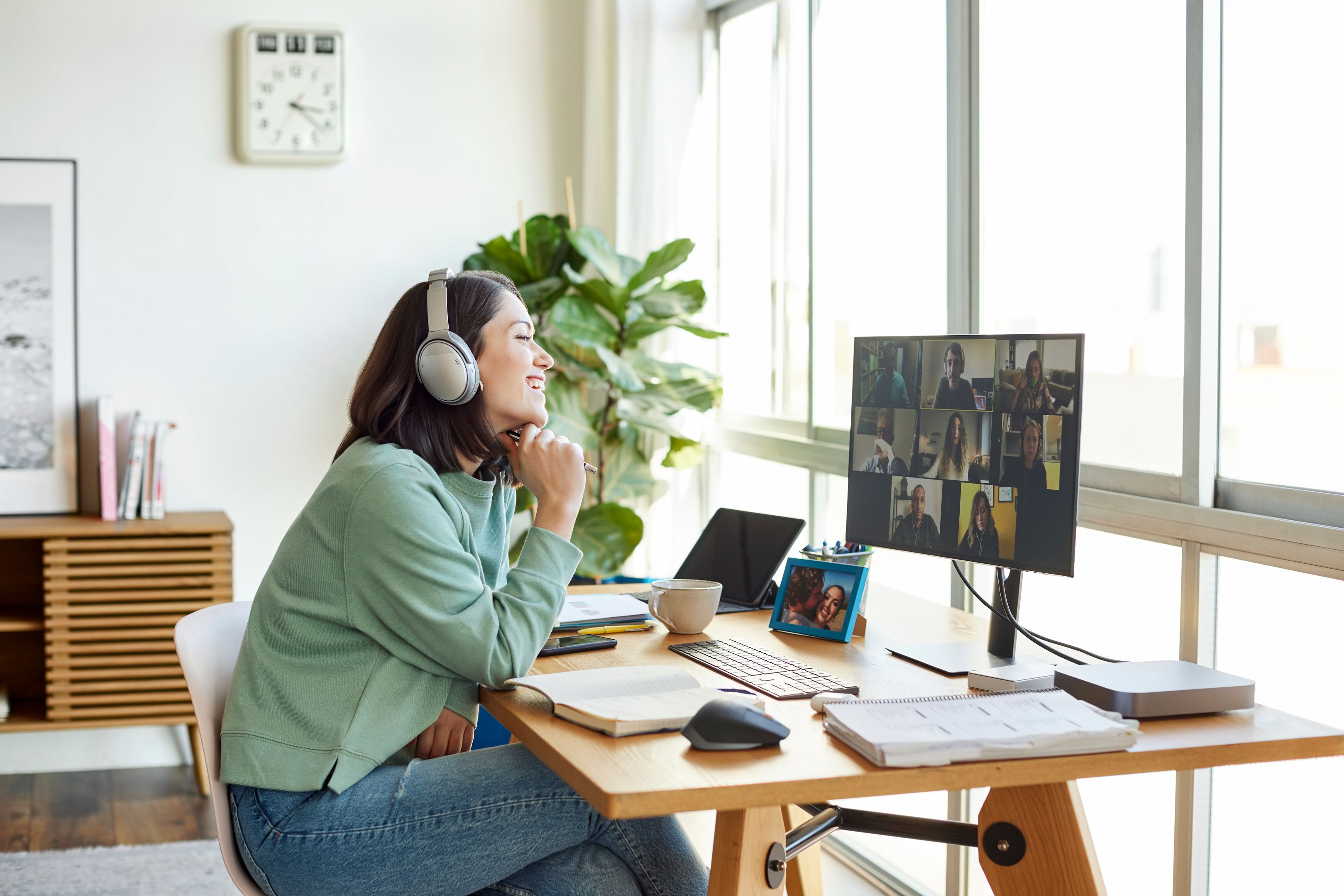 Person wearing headphones and smiling while participating in a video conference on a computer, with multiple monitors displaying various work-related windows.