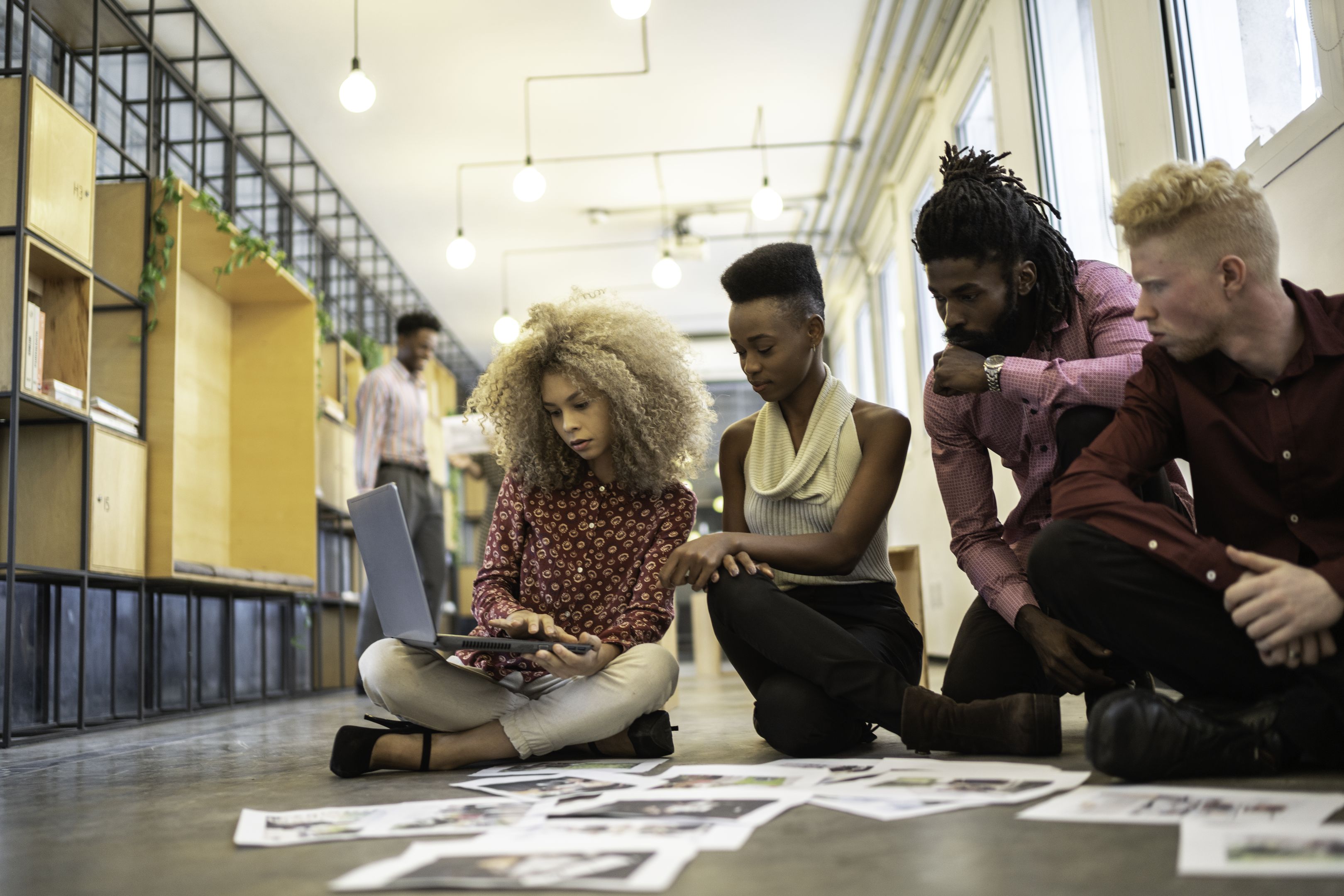 A group of people sitting on the floor reviewing images on a laptop and scattered papers in a modern office space.