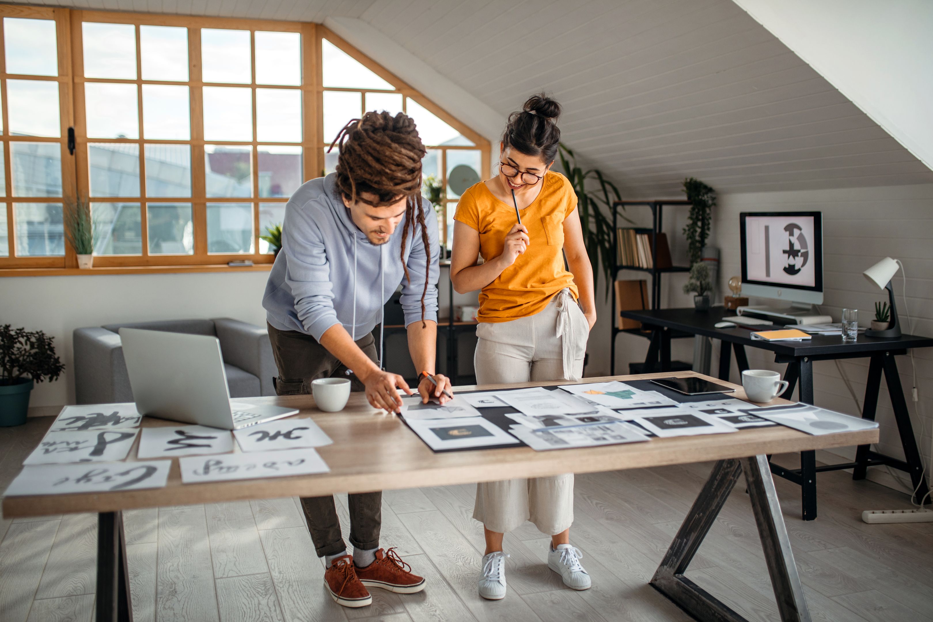 Two people review designs and photographs spread out on a table in an office space.