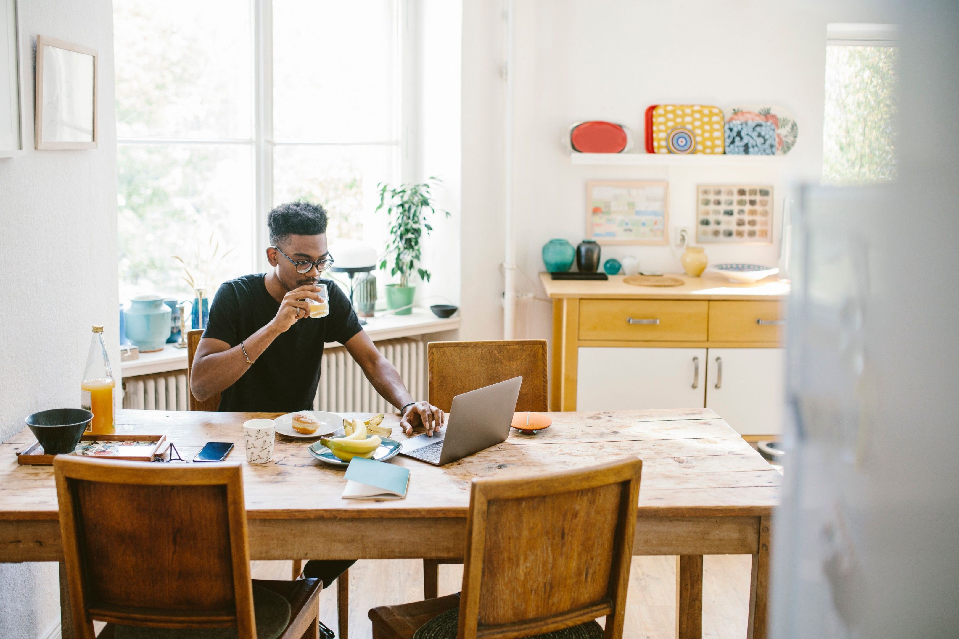 Person works from home on a laptop while having breakfast at a wooden table in a bright, cozy kitchen.