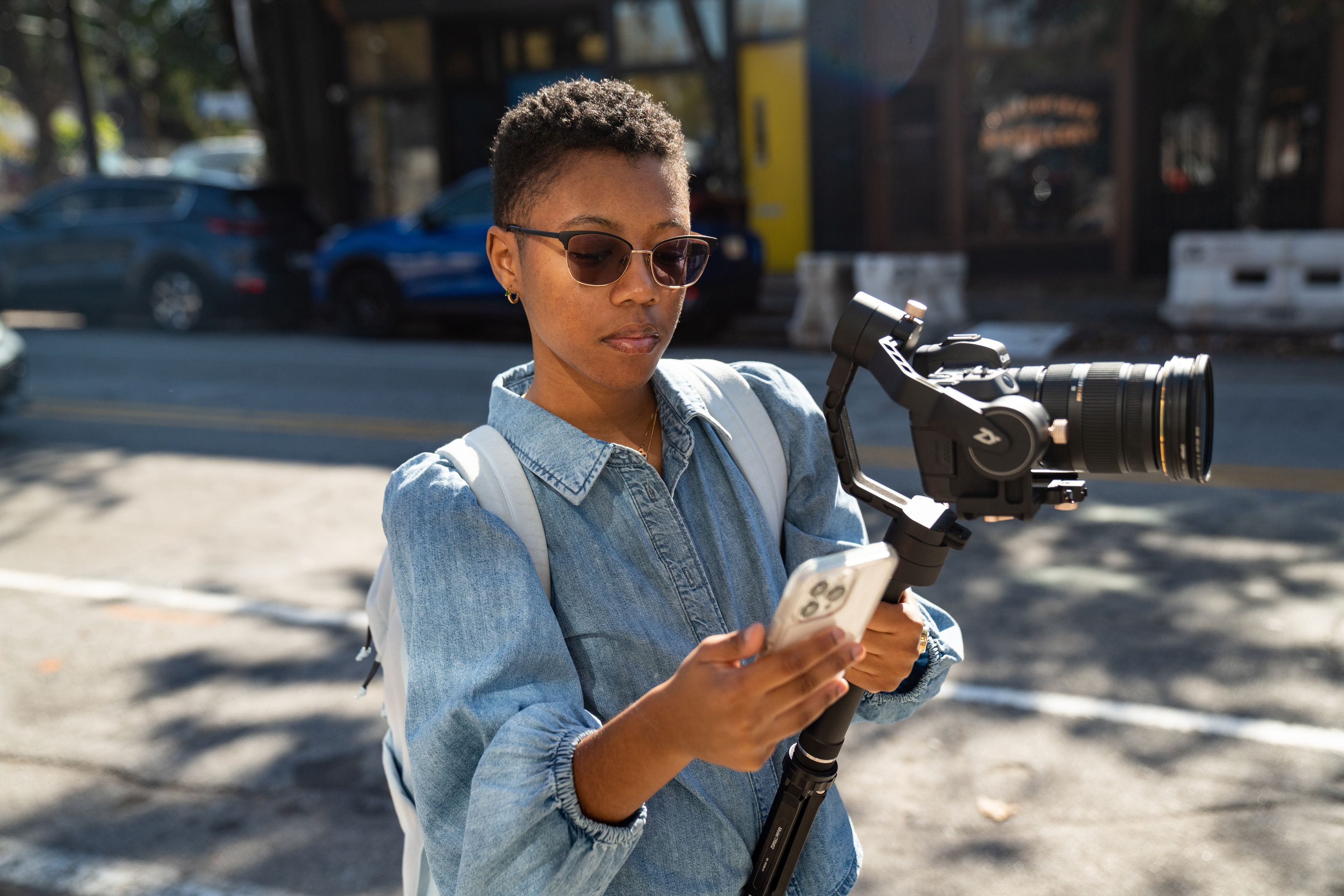 Person holding a camera stabilizer and checking a smartphone while standing on a street with parked cars in the background.