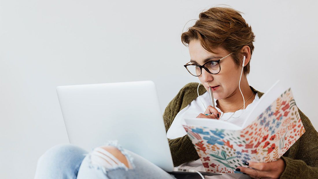 A person wearing earphones working on a MacBook and holding a notebook and pen.