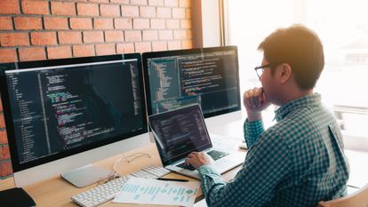 A man with glasses working on a laptop with two large computer monitors behind it