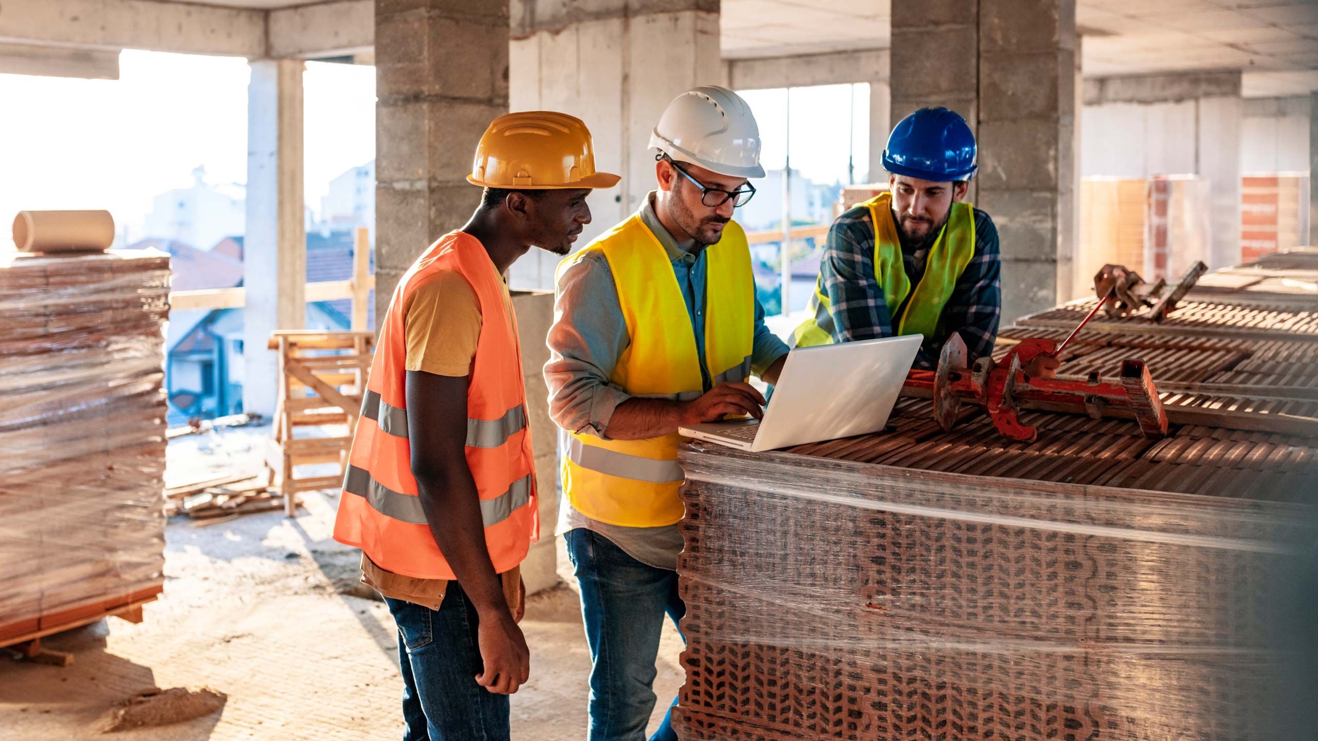 Three construction workers wearing hard hats and safety vests looking at a laptop