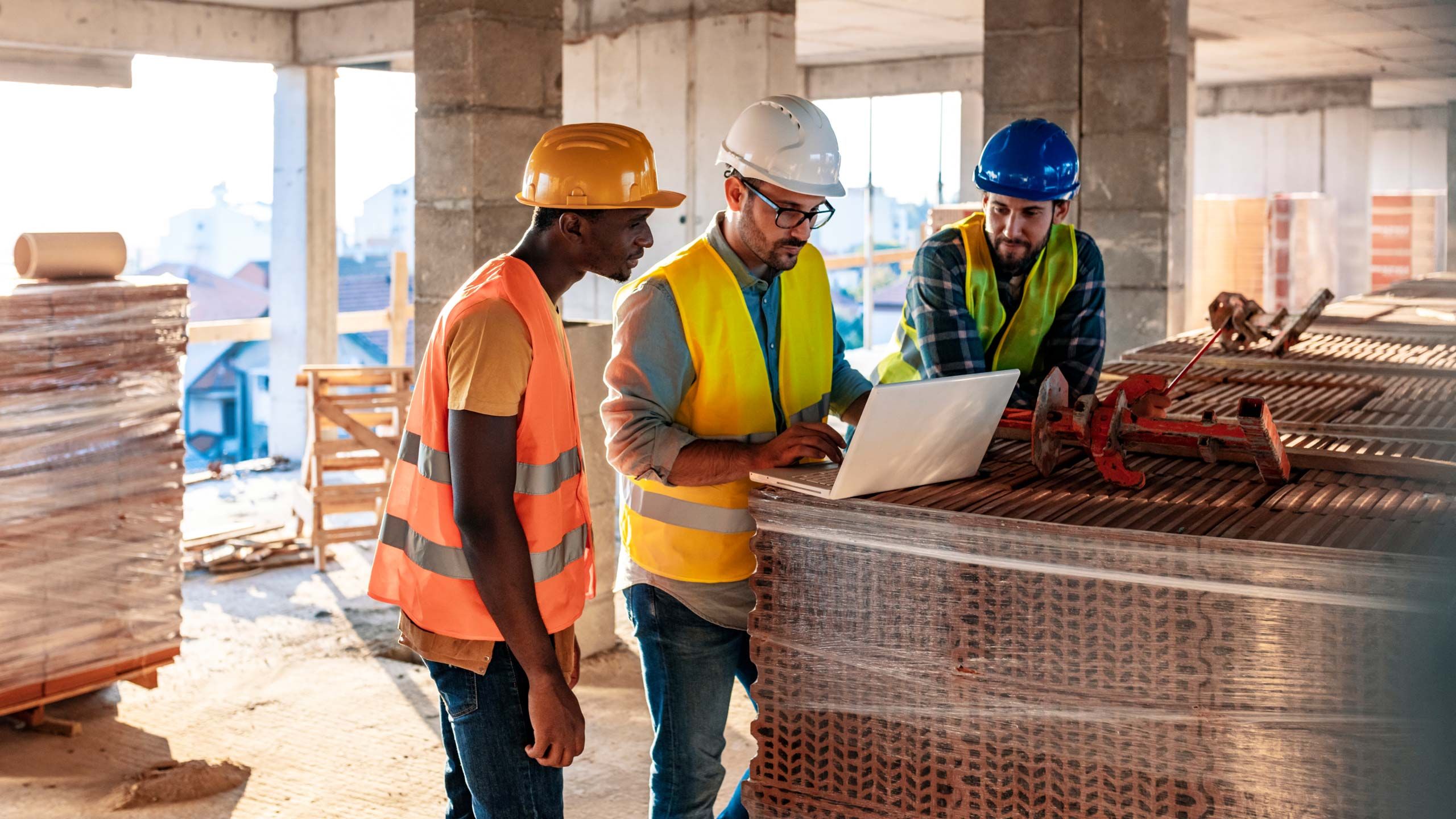Trois ouvriers du bâtiment portant des casques de chantier et des gilets de sécurité qui regardent un ordinateur portable