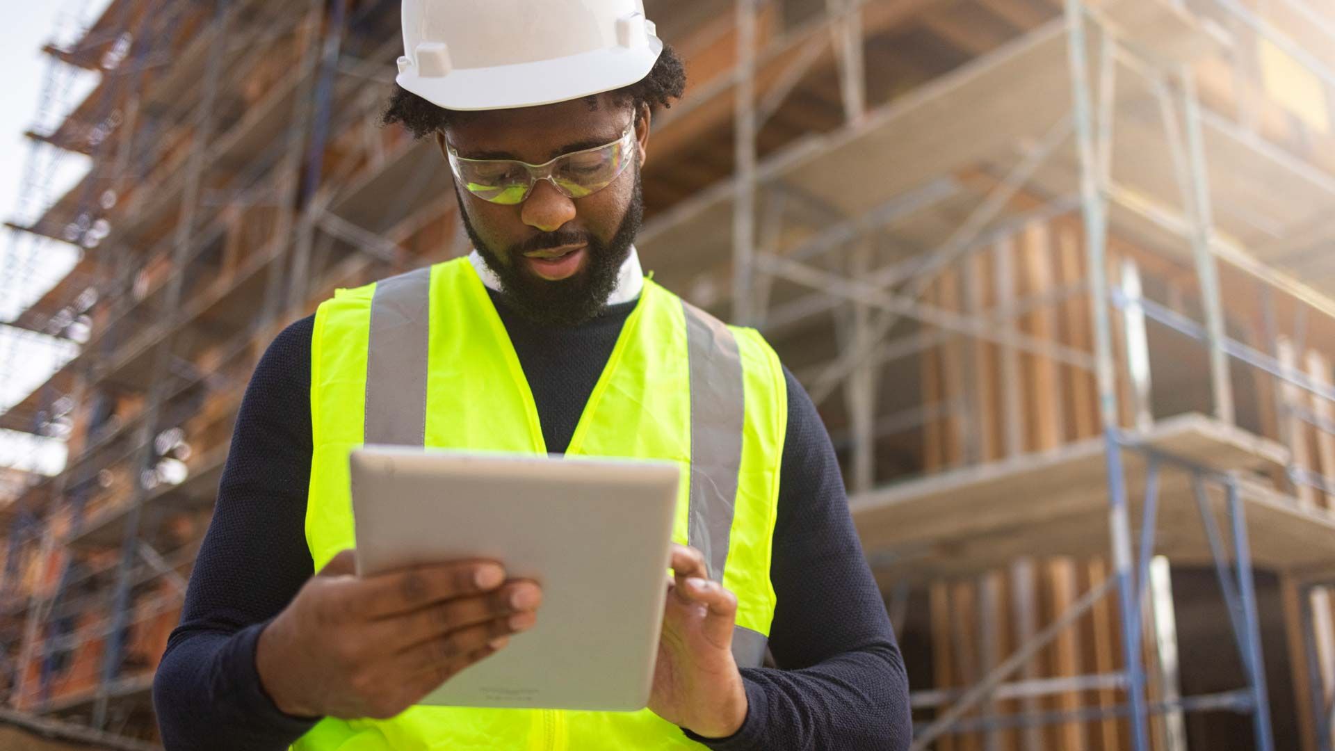 A man wearing a hard hat, safety goggles and safety vest, standing in front of a building under construction