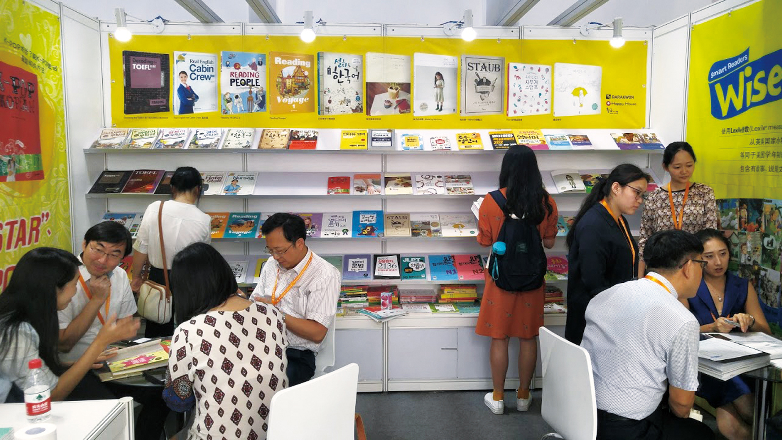 People sitting at tables and standing looking at books and learning material with vibrant yellow walls around them