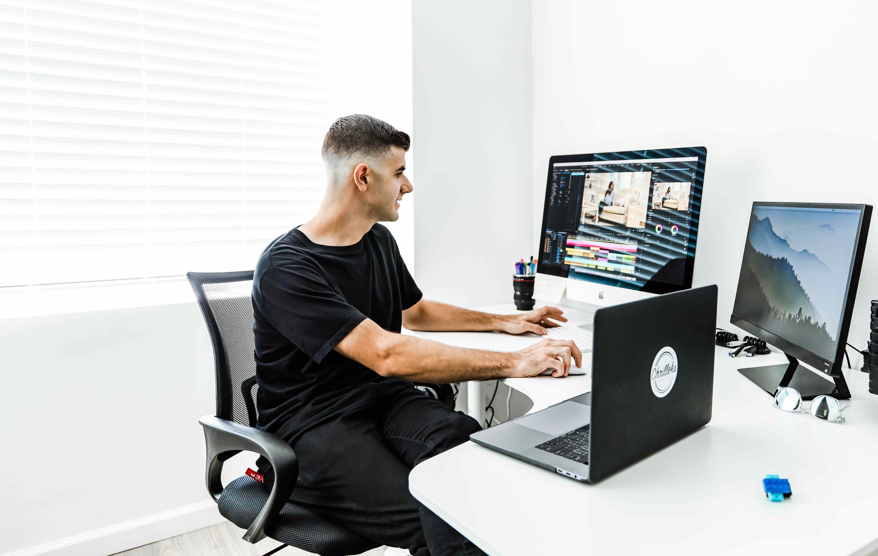 Chris Grubisa sitting at a white desk with two monitors and a laptop editing video