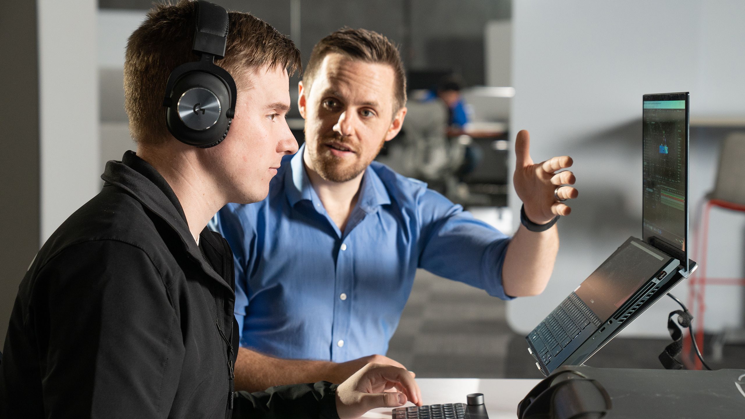 Man in blue shirt talking to man wearing a black shirt and black headphones while looking at a laptop