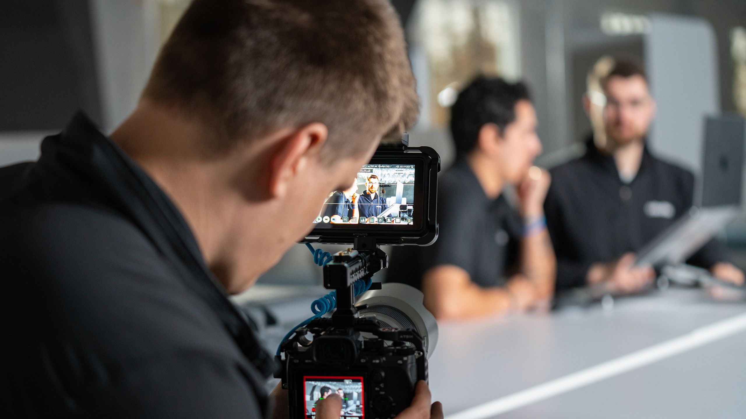 Up close of the back of a man’s head as he’s looking through a view finder on a camera setting up a shot of two men talking at a table.