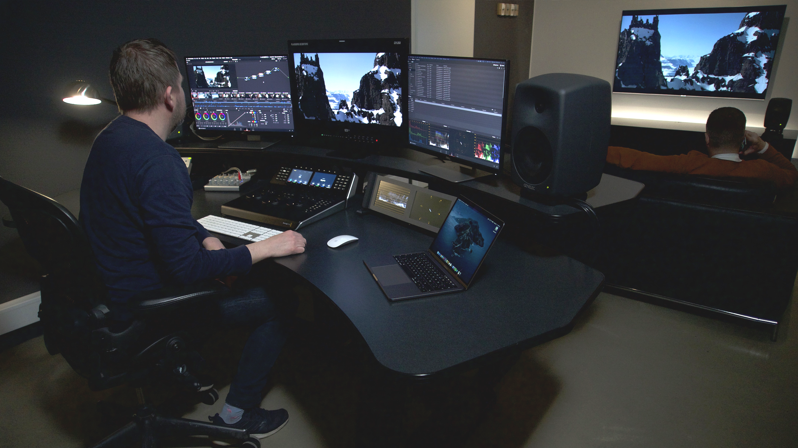 Man working at an editing suite with three monitors at a black desk and a laptop. 