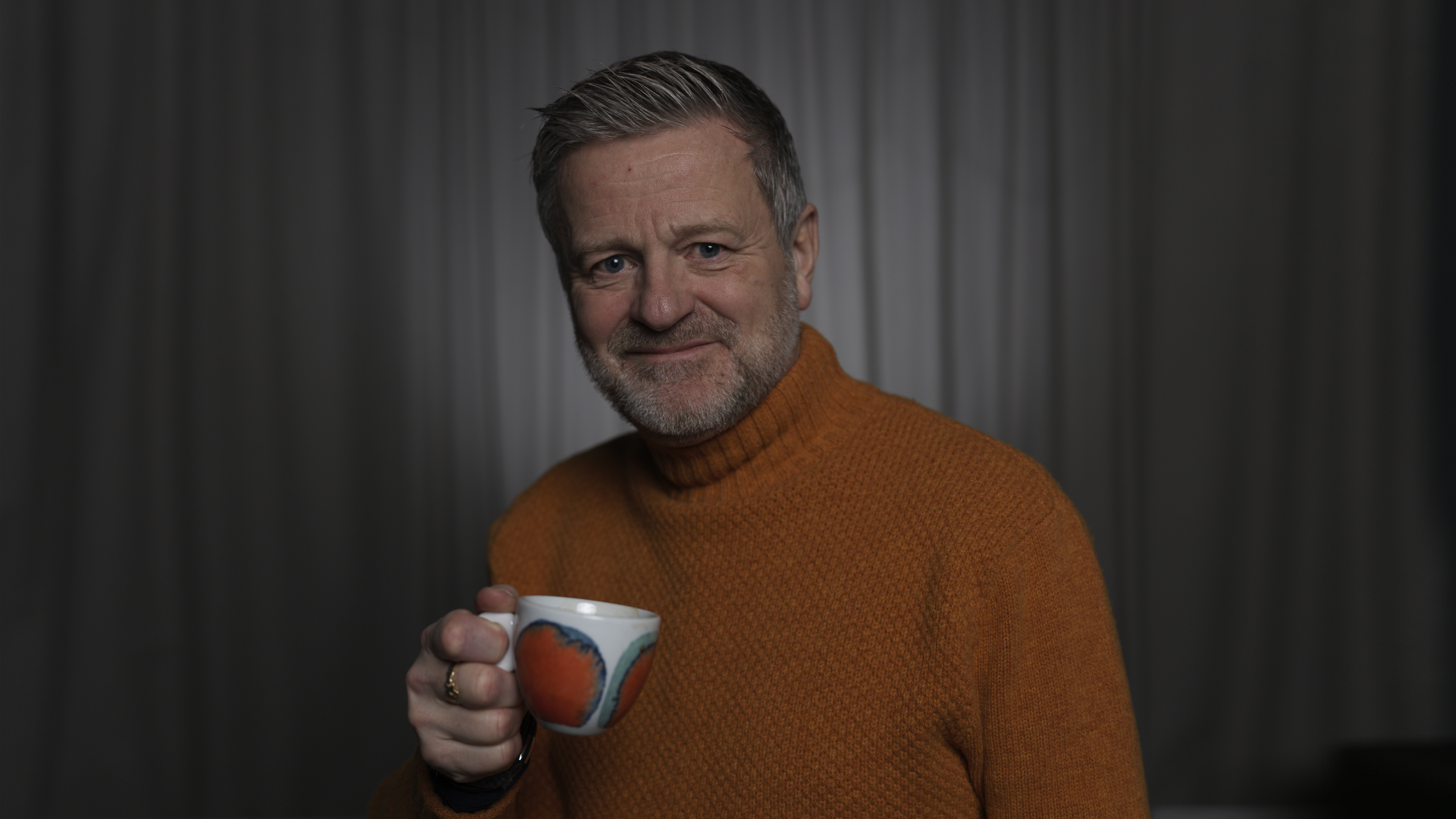 Örn Sveinson in an orange turtleneck holding a tea cup in front of a gray wall