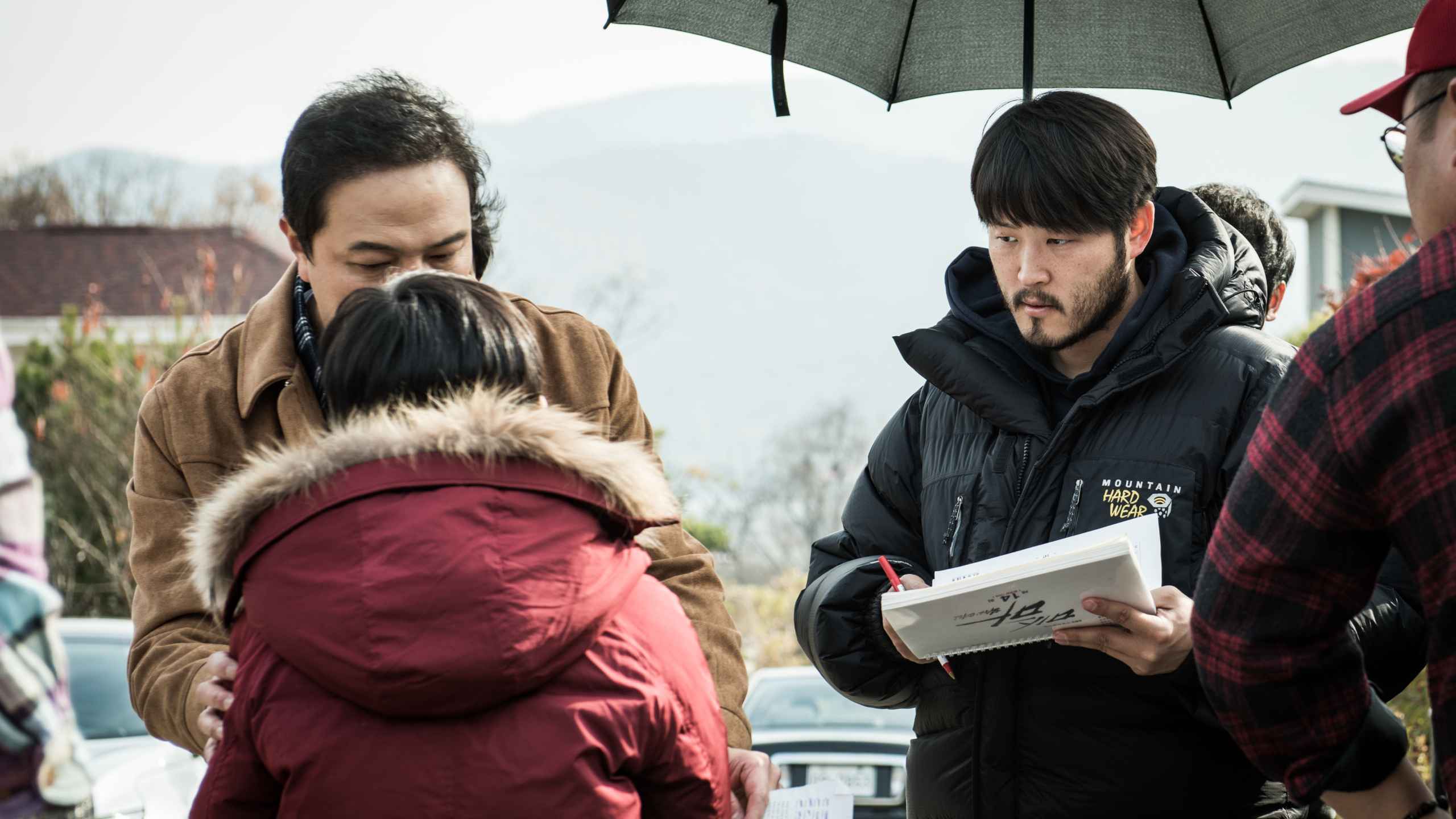 Yun Hong Min standing in a large coat under an umbrella making notes on a script while listening to two other speak