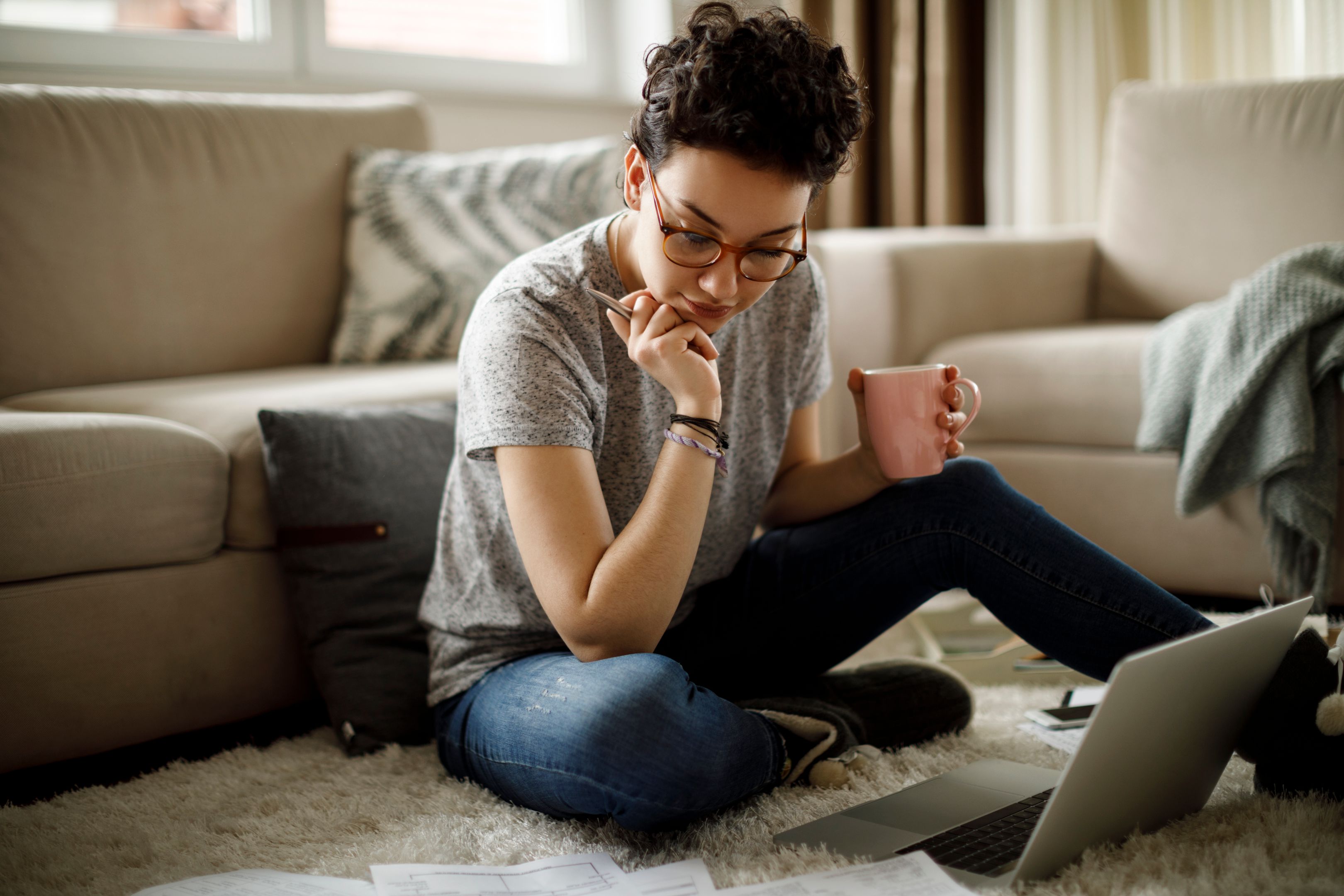A person holding a pen and mug works from their laptop on the floor of their home.