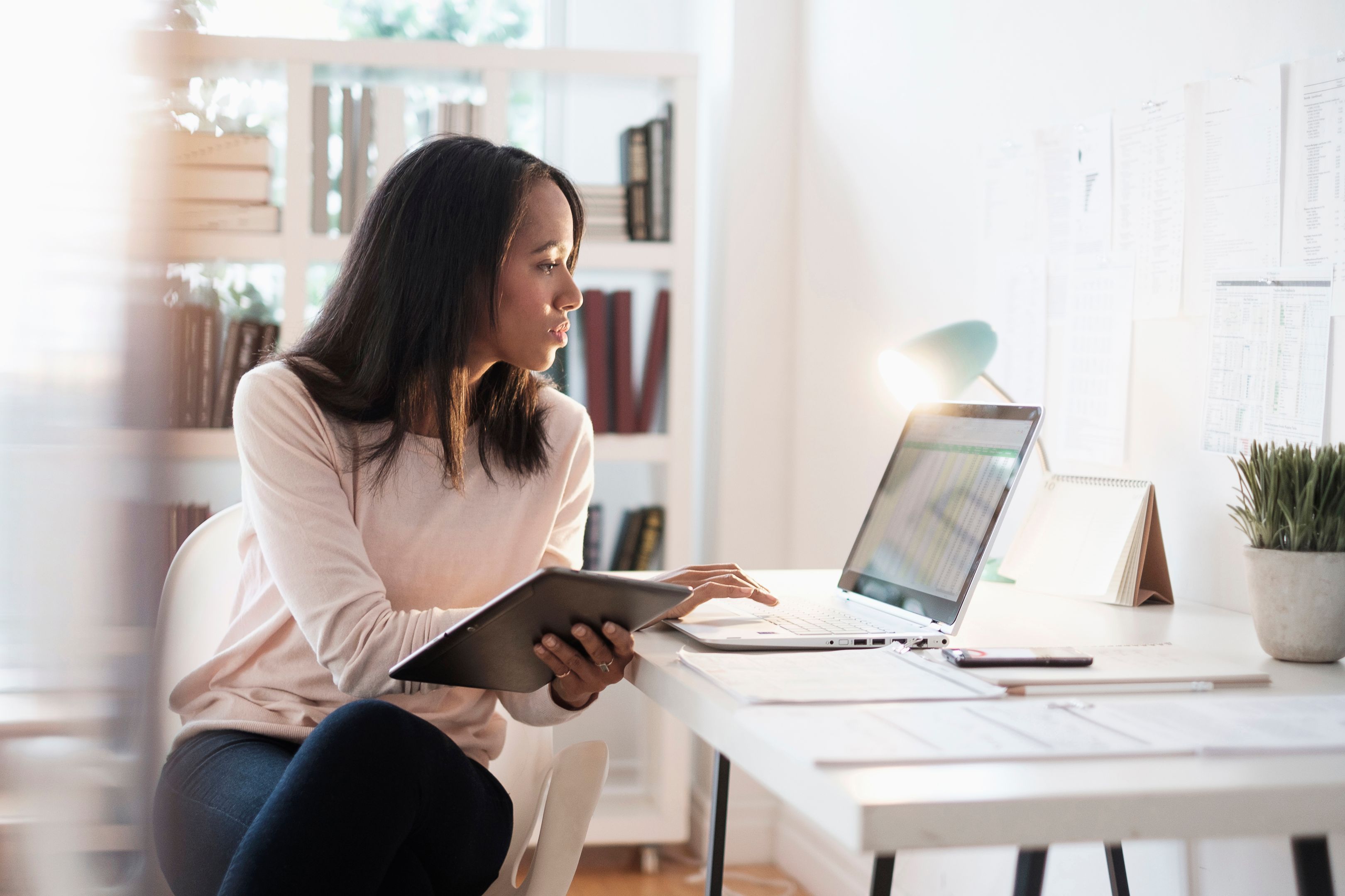 A person working on a laptop and tablet in a home office with a desk lamp.