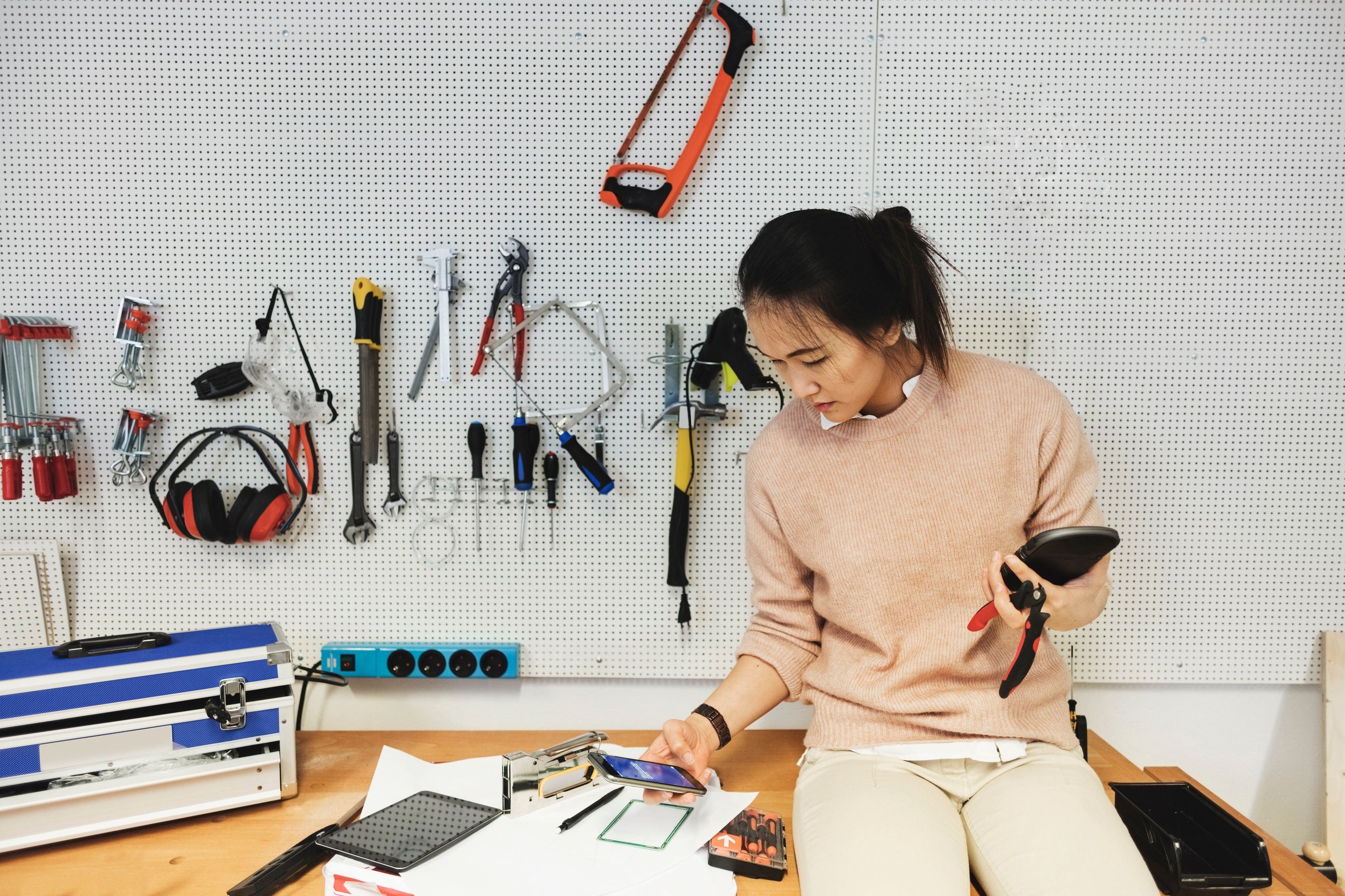 A person working with tools in a workshop holding their mobile device.