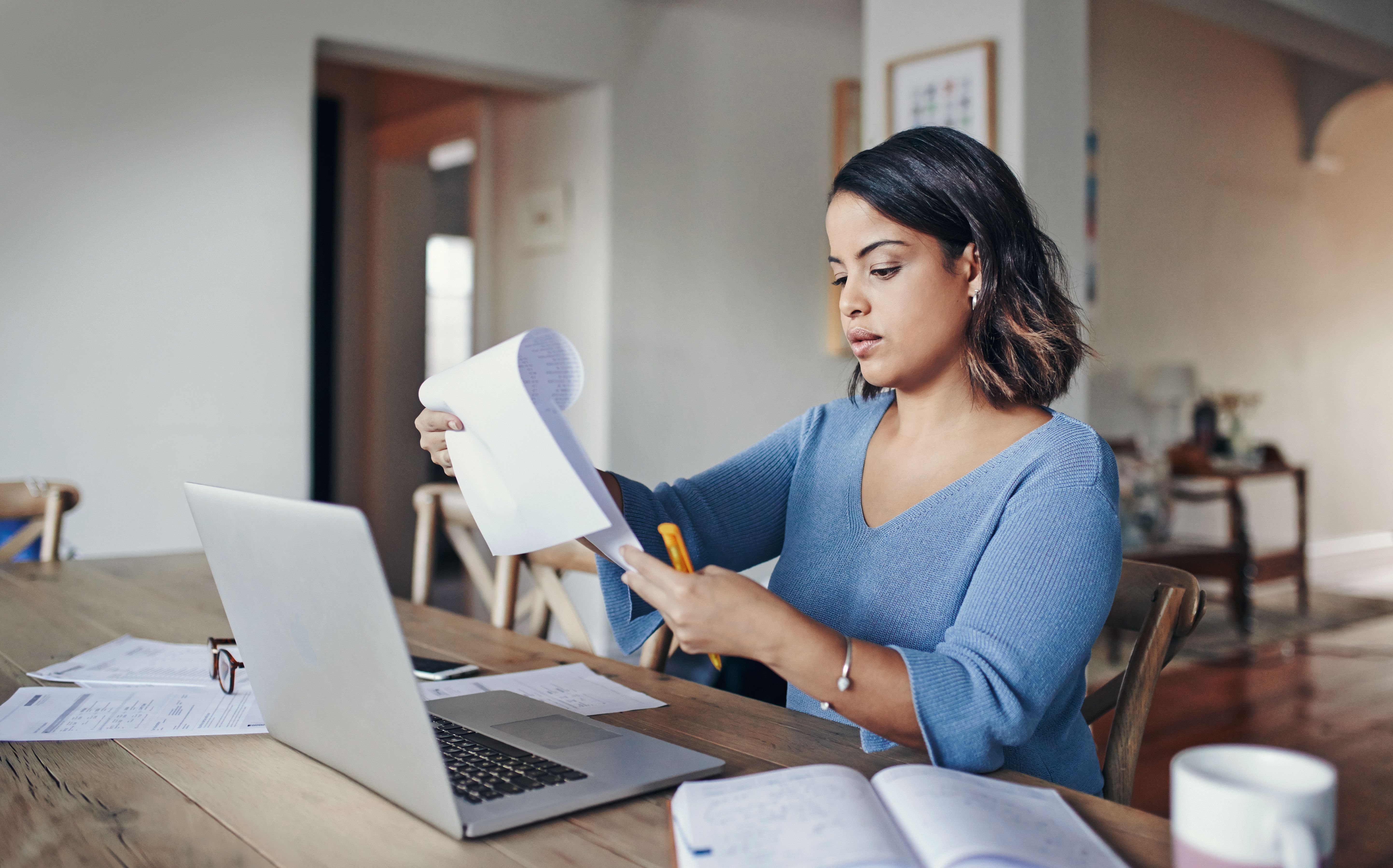 A person holds a pen while looking through a paper documents in front of a laptop at home.