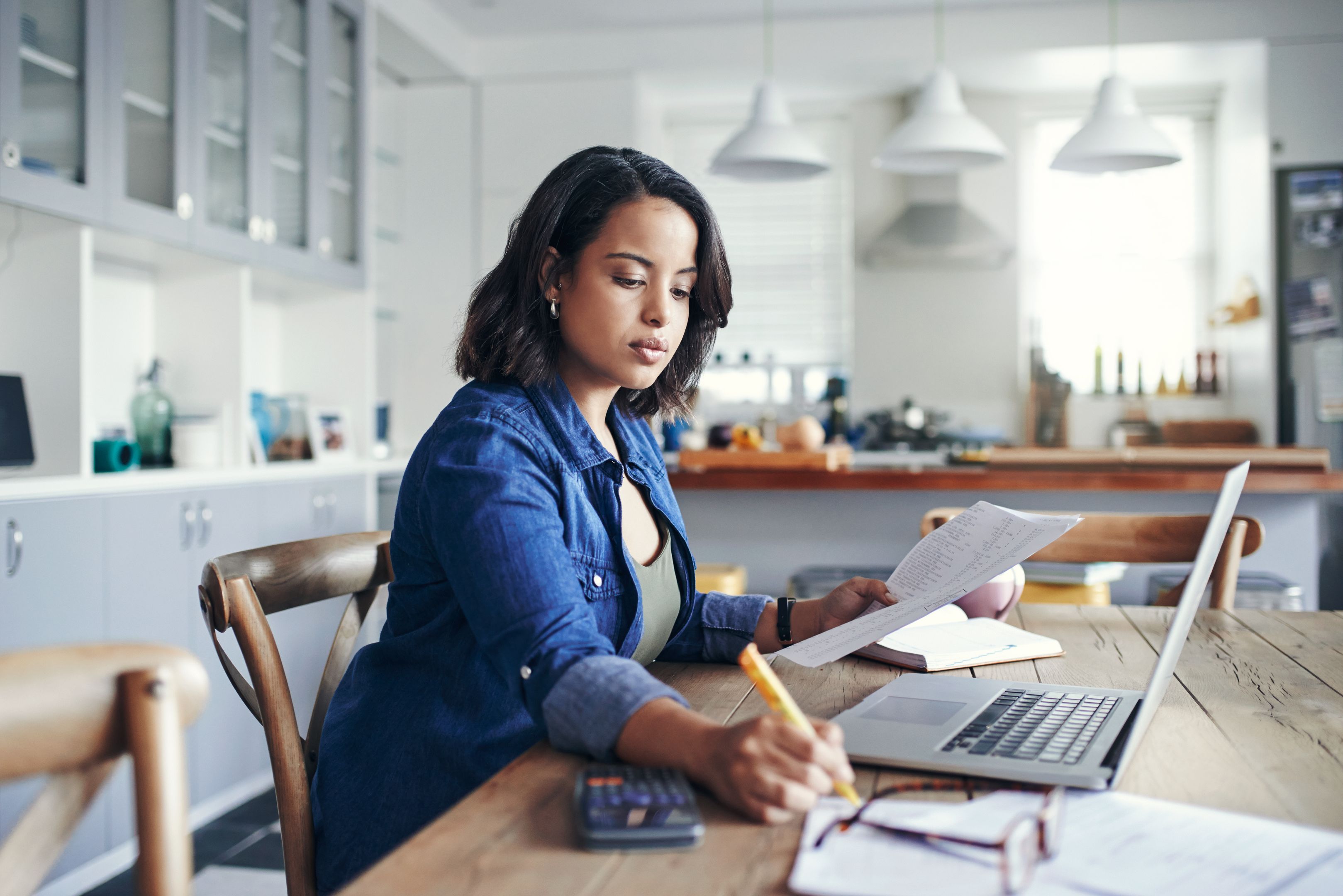 A person is making notes while holding papers and sitting in front of a laptop.