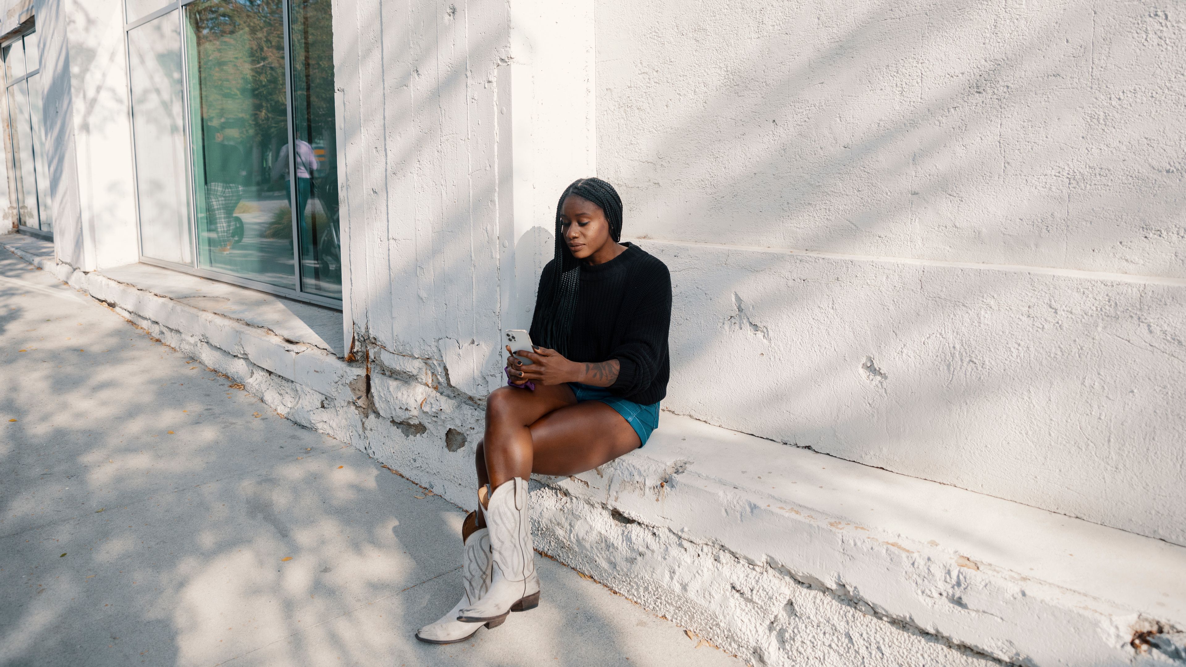 A person sitting against a wall outdoors looking at their mobile device.
