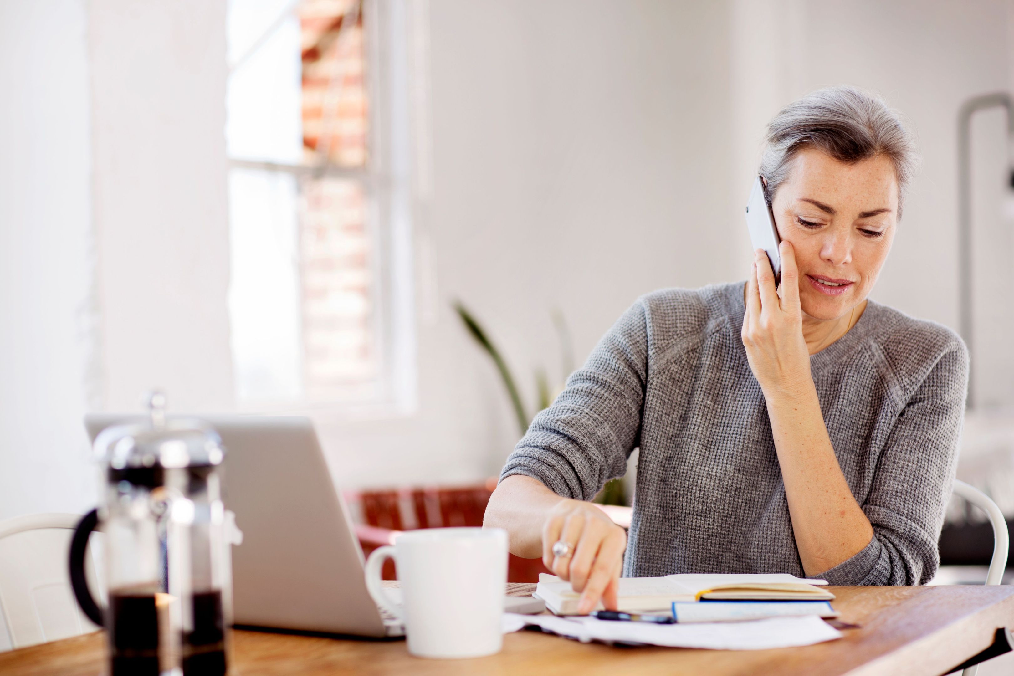 A person working on a laptop from home while taking a call on their cell.