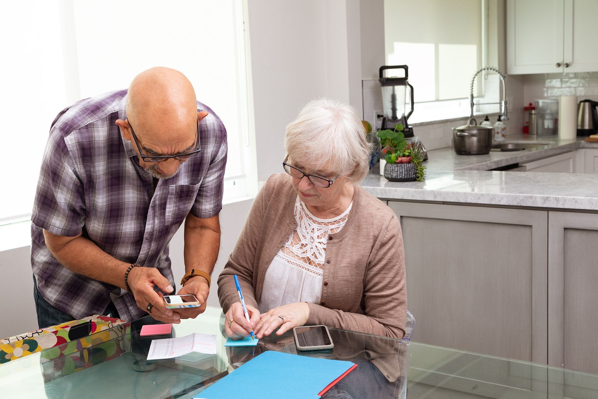 An older couple organize their plans, with one reading from their mobile device as the other makes notes on Post-Its.