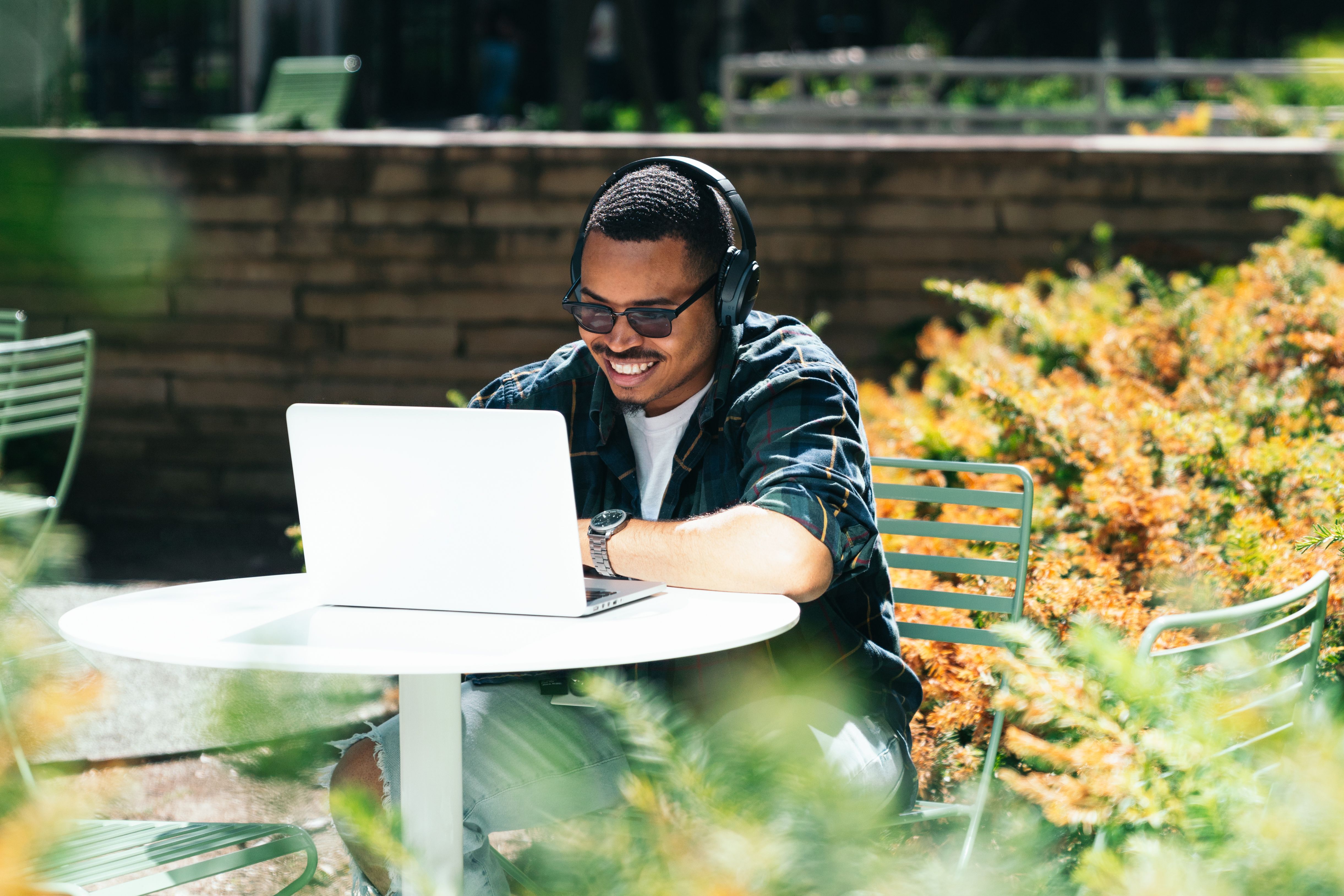 A person wearing headphones and sunglasses sat on table outside, working on their laptop.