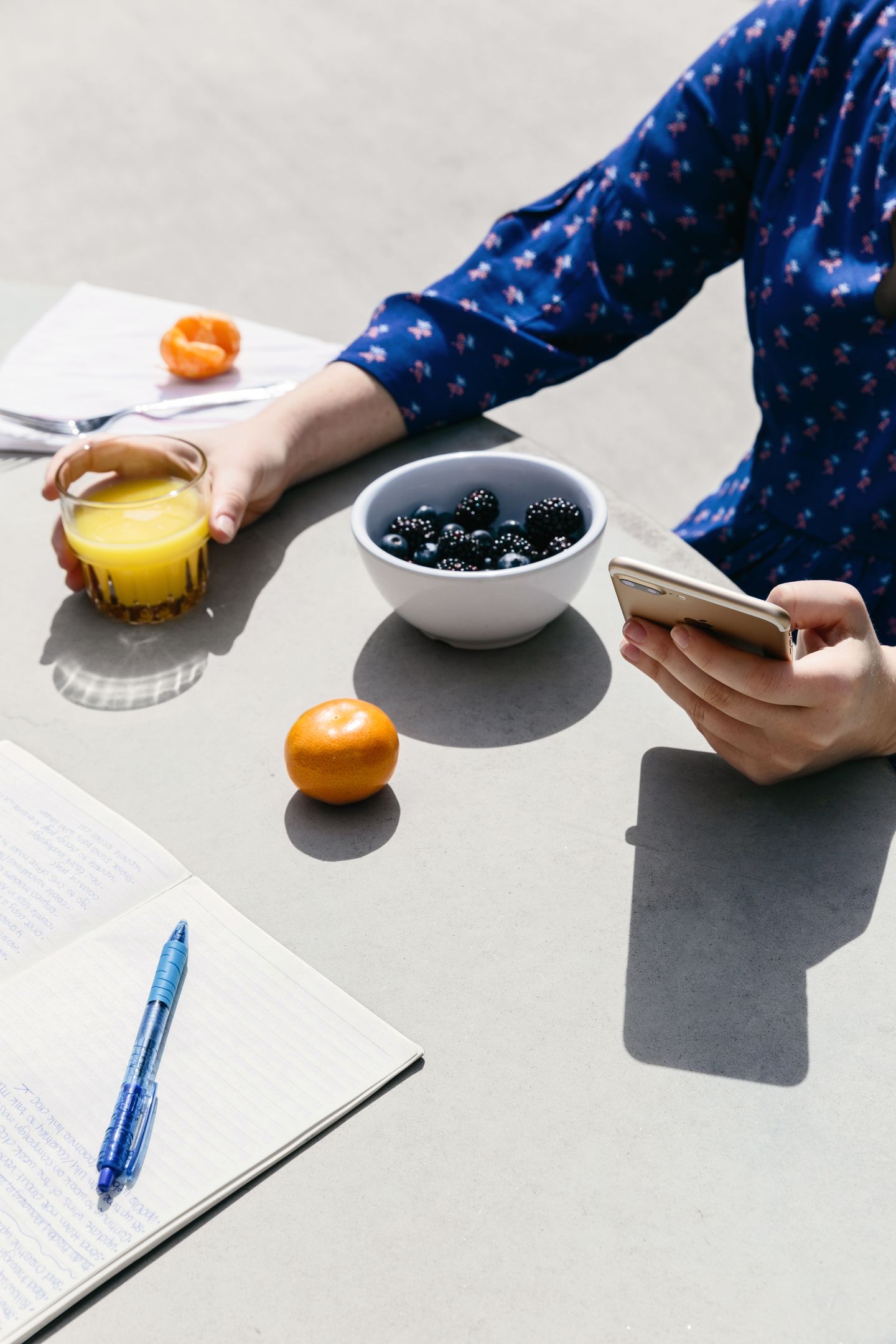 A person sitting at a table outside on their mobile device while holding a glass of orange juice.