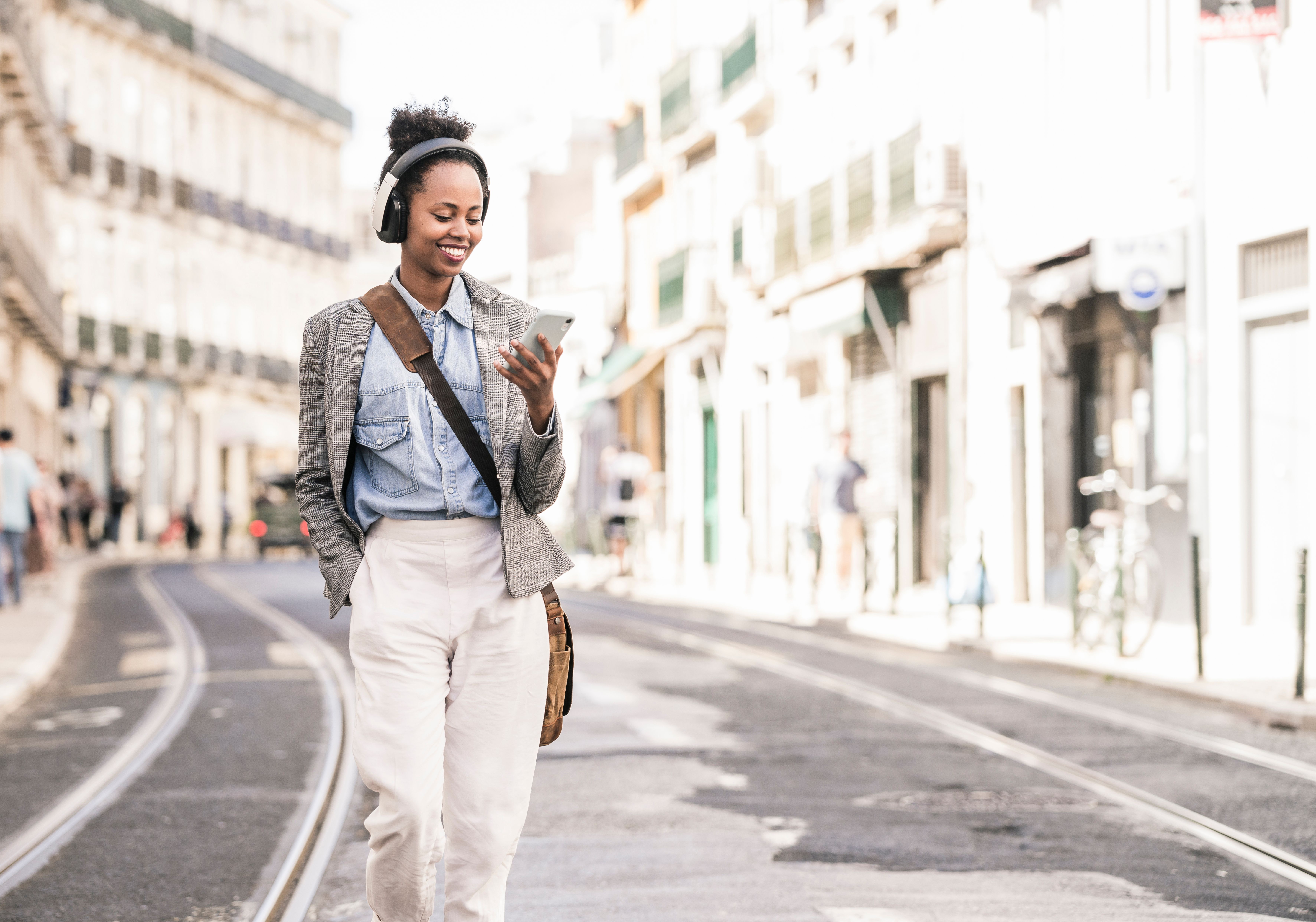 A person wearing headphones smiles at their mobile device as they walk down a road.