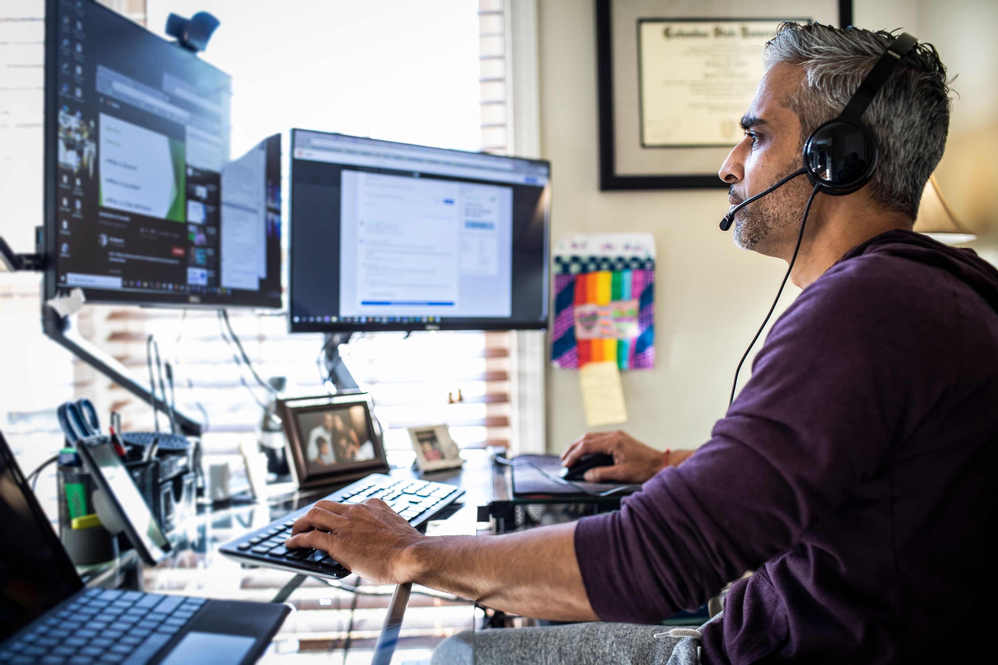 A person wearing a headset works on two monitors from a home office.