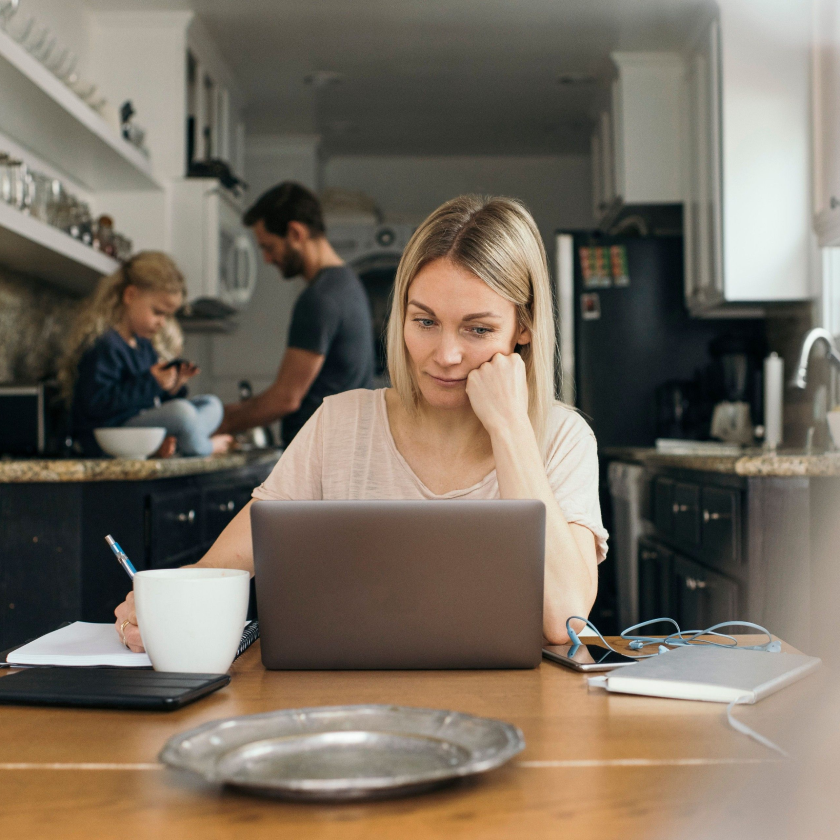 Person working on a laptop at a kitchen table, with family in the background, illustrating a work-from-home setting.