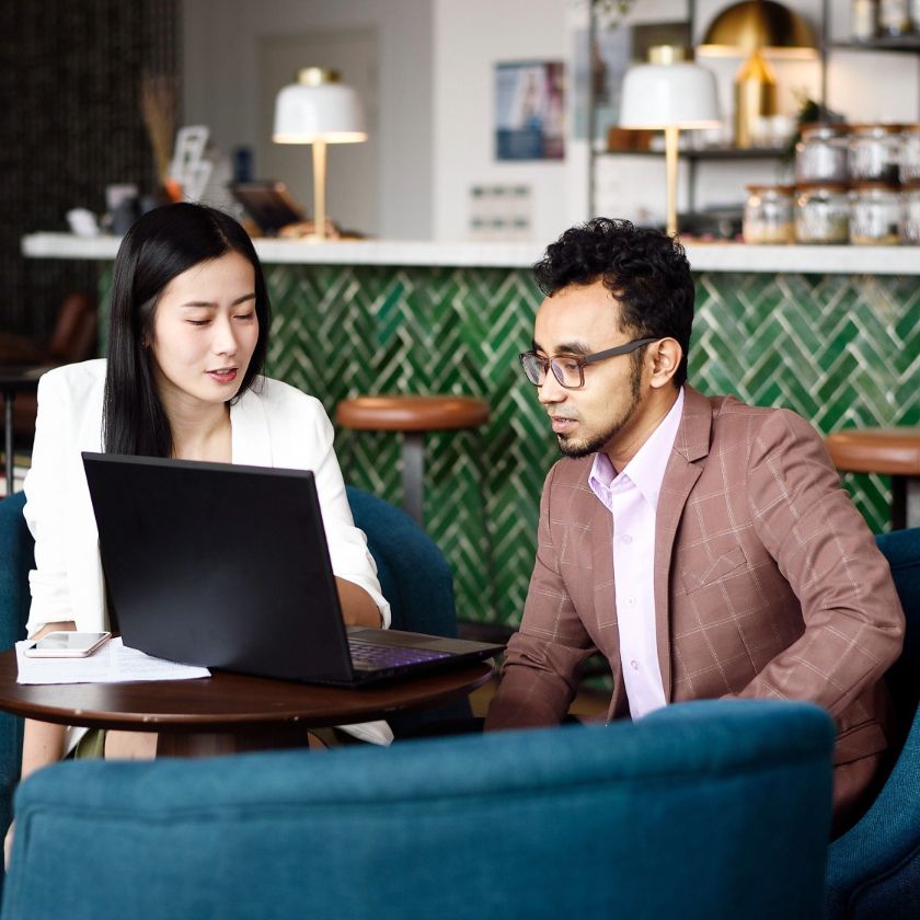 Two sales people sat in a cafe discuss a sales opportunity while looking at a laptop on a table.