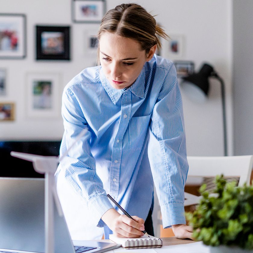A woman stands in front of her laptop, taking notes down on a notepad