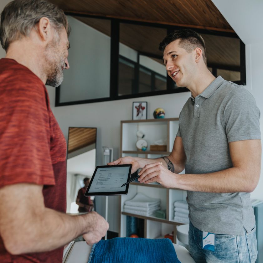 A salesperson holds out a tablet device to a customer, pointing to the screen with a stylus to show where to sign a document.