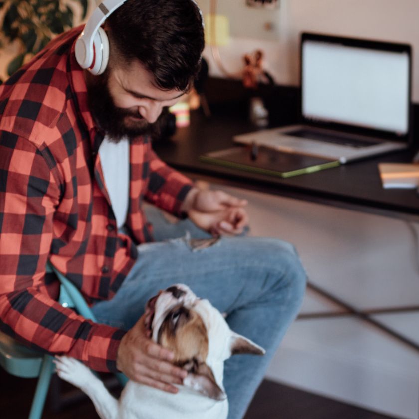 A man sitting by his laptop at a desk whilst working from home bends down to pet his dog