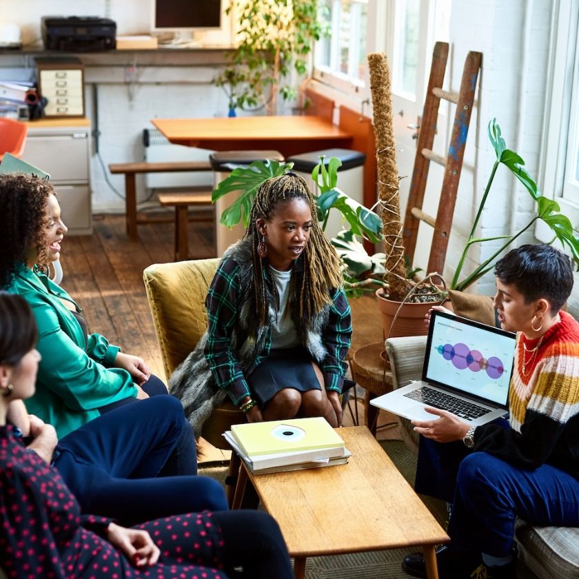 A group of people sit in an office breakout area discussing an issue around a laptop.