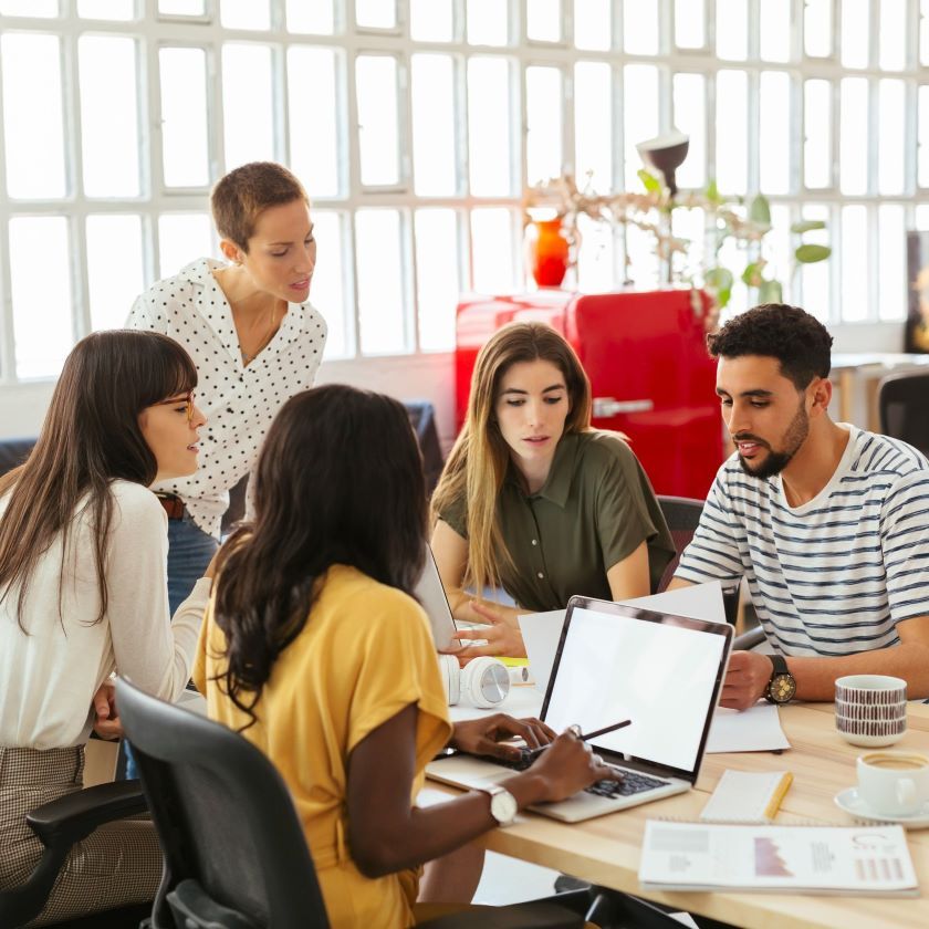 A creative team collaborates around a laptop in a modern office, discussing ideas that may involve or be impacted by AI.