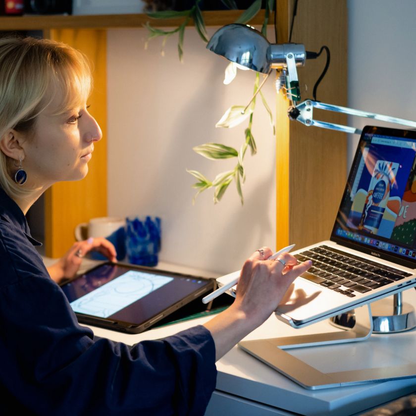 Person sitting at desk typing on computer with stylus in hand, tablet in background.