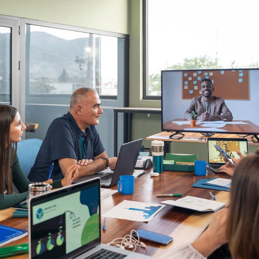 Group of people in an office run a brainstorming session on a video call with a remote colleague displayed on a large screen.