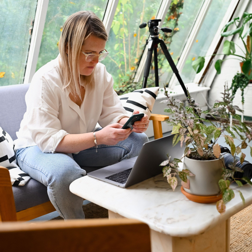 En person sitter på en sofa foran en bærbar datamaskin og bruker en mobilenhet for å sikkerhetskopiere de nyeste bildene sine til skyen.