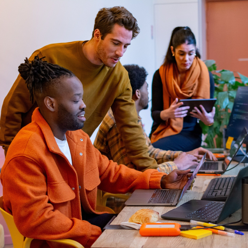 Four coworkers—two sitting, two standing—collaborate on laptops and tablets for a project.