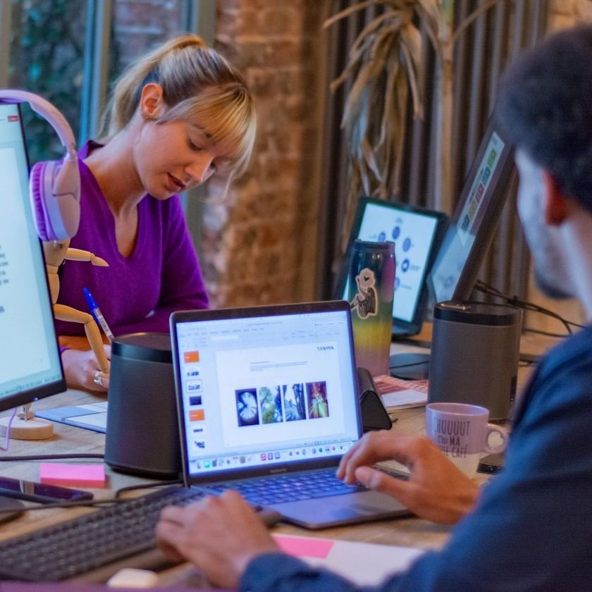Two people work at a desk with laptops and monitors, one person is working on slides, and the other is writing.