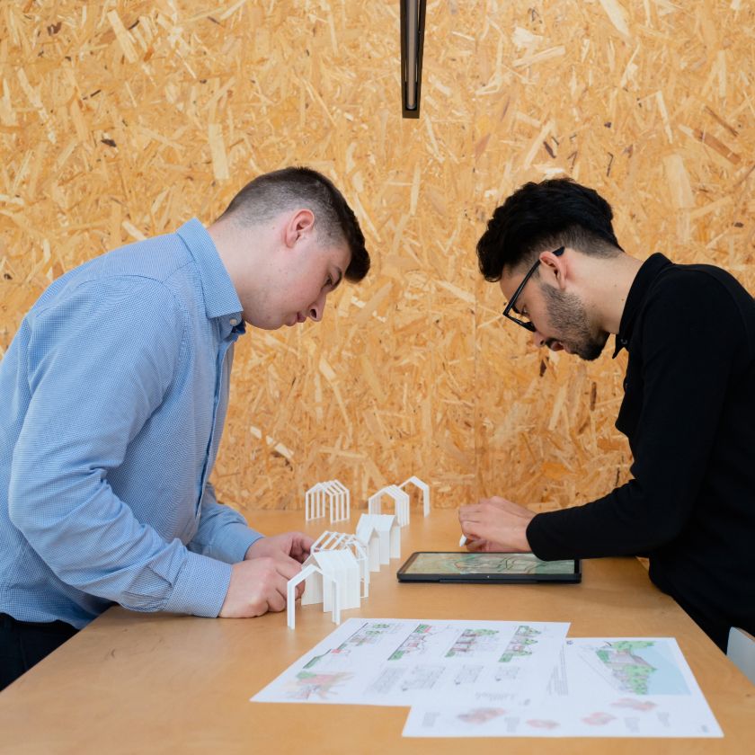 Two architects consult a design file on a tablet device while working with scale models at a tabletop.