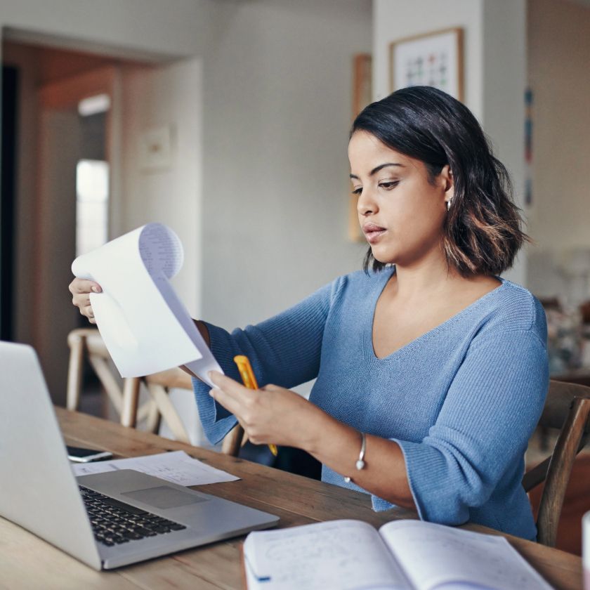 A person sits writing at their laptop on a desk covered in books, papers, and pens.
